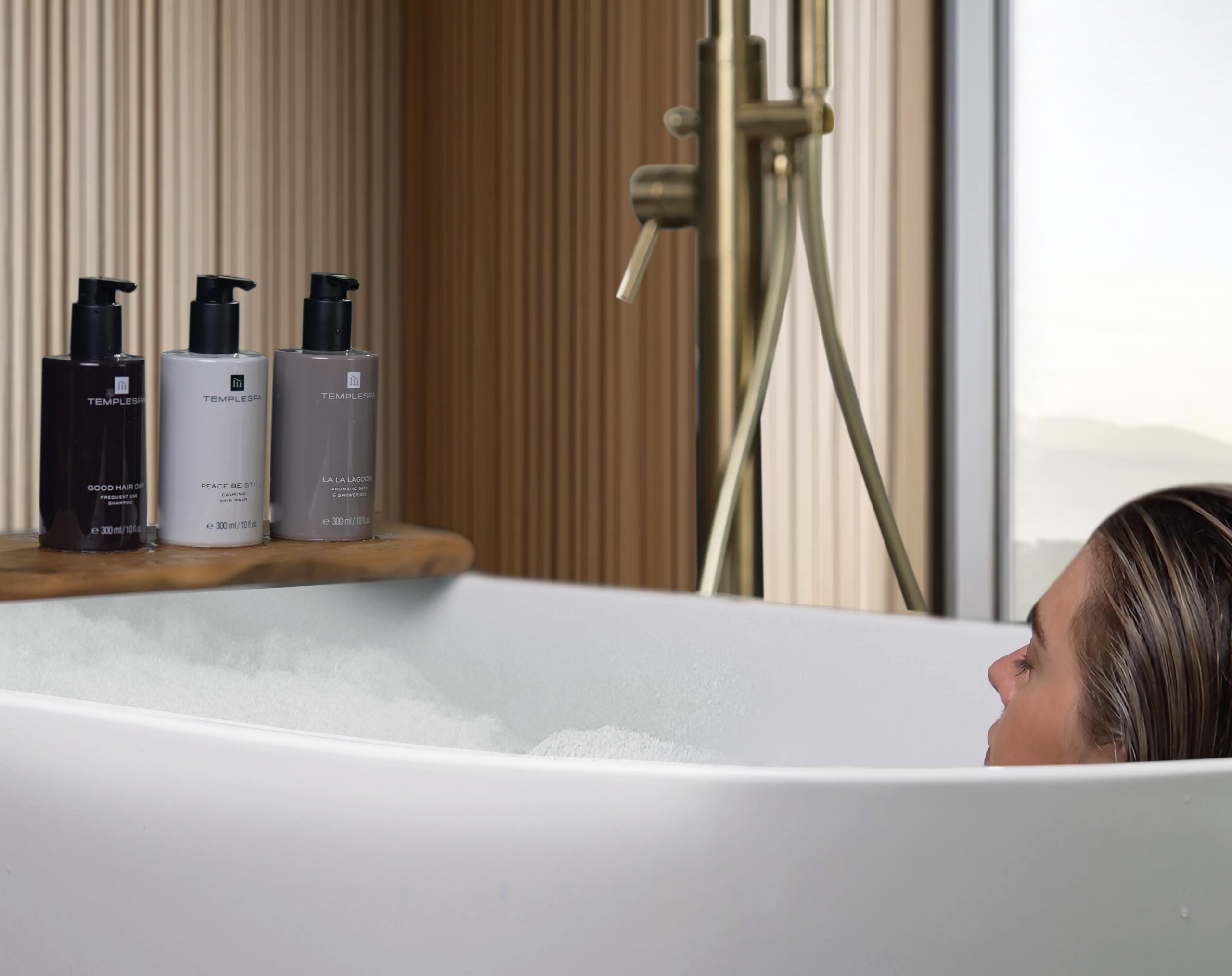 Lady in bath tub with lots of bubbles, with TEMPLESPA products showing, LA LA LAGOON shower gel, PEACE BE STILL body moisturiser and GOOD HAIR DAY shampoo 