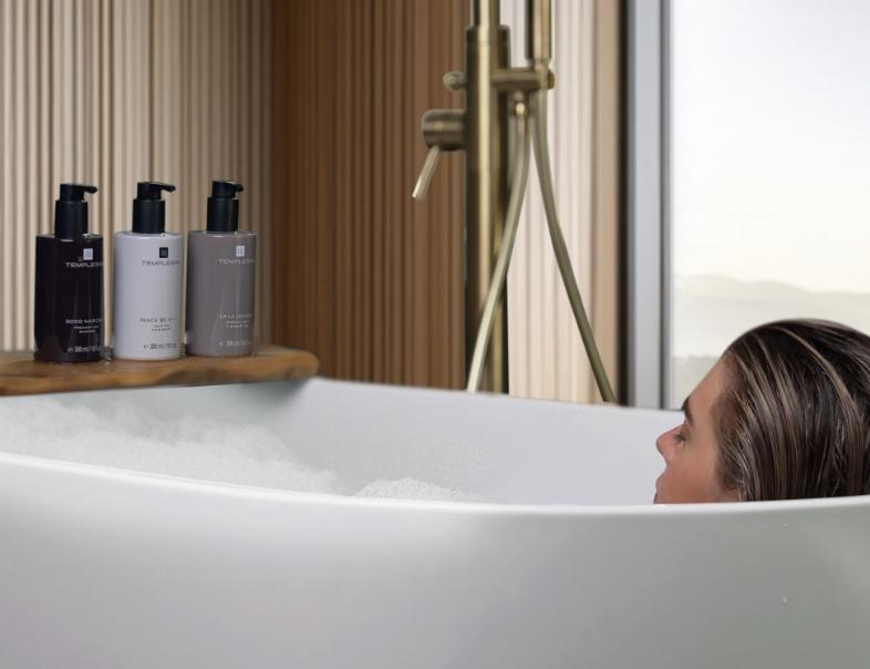 Lady in bath tub with lots of bubbles, with TEMPLESPA products showing, LA LA LAGOON shower gel, PEACE BE STILL body moisturiser and GOOD HAIR DAY shampoo 